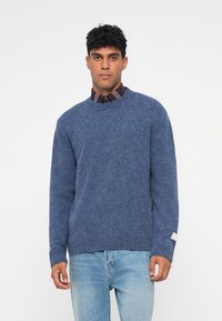 Maglione blu lavorato a maglia con scollatura a girocollo, polsini e orlo a costine, indossato sopra una camicia a quadri, completato da un'etichetta sulla manica.
