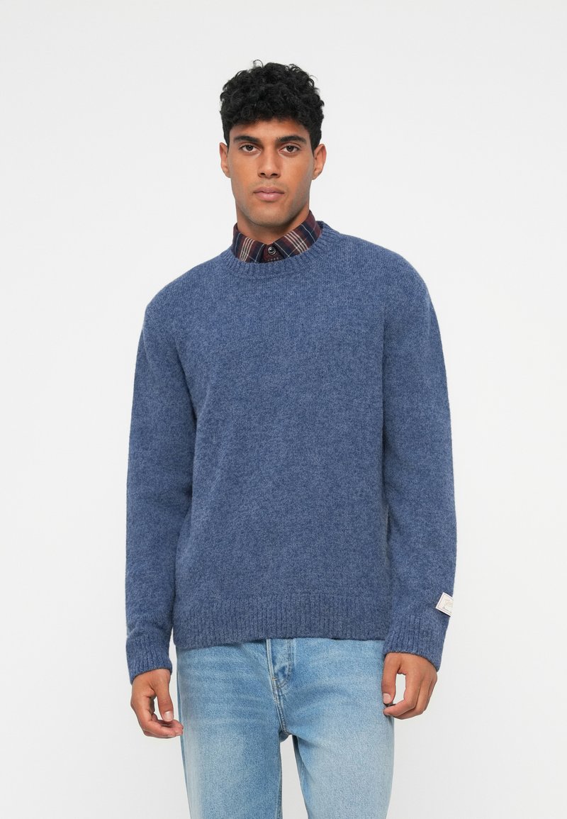 Blauwe gebreide trui met een ronde hals, geribbelde manchetten en zoom, gedragen over een geruite blouse, aangevuld met een label op de mouw.
