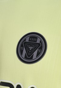 Neon guldt stof med en tekstureret overflade, der har et fremtrædende sort cirkulært emblem. Emblemet inkluderer et trekantet design og "XXIII."