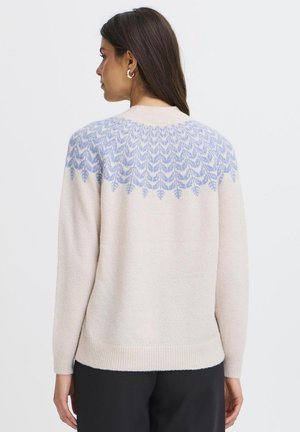 Vrouw draagt een crèmekleurige trui met een blauw gebreid patroon over de bovenrug, gecombineerd met zwarte broek, met de rug naar de camera op een effen achtergrond.