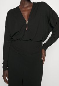 Black long-sleeve top with a deep V-neck and a decorative gold button at the center. Smooth fabric with a slightly loose fit and gathered detail.