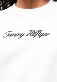 Valkoinen huppari, jossa on näkyvä kirjailtu "Tommy Hilfiger" -logo mustalla kaunokirjoituksella. Pehmeä pinta klassisella pyöreällä kaula-aukolle.