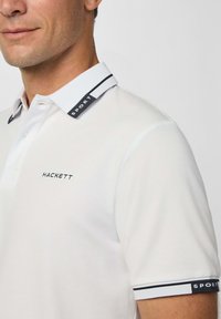 Muž v bílém polo tričku s vyšitou značkou "HACKETT" na hrudi a nápisem "SPORT" na límci a manžetách.