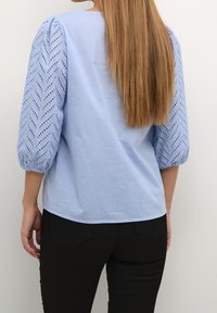 Ljusblå blus med puffiga, mönstrade ärmar som har en chevron-design och utskärningar. Bakifrån ses en avslappnad passform och slät textur.