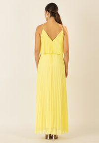 Gelbes, plissiertes Maxikleid mit V-Ausschnitt, verstellbaren Spaghetti-Trägern und fließender Silhouette, kombiniert mit Riemchenschuhen.