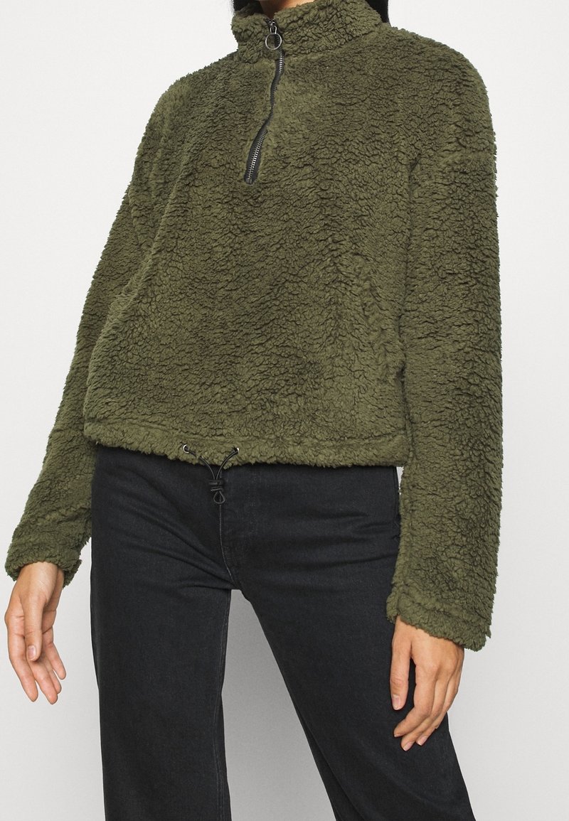 Sudadera de forro polar verde oliva con cuello alto, cierre de cremallera y dobladillo con cordón ajustable. Textura suave y ajuste relajado con mangas largas.