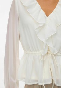 Hvit bluse med rysjet hals, lange semi-gjennomsiktige ermer, elastisk midje med snor, og peplum-bunn.