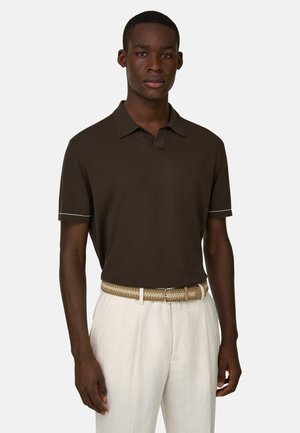 Uomo che indossa una polo a maniche corte marrone scuro, cintura intrecciata beige e pantaloni chiari, in piedi davanti a uno sfondo neutro.