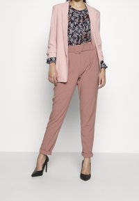 Žena v růžovém saku, květinové blůze, růžových kalhotách s páskem a ohrnutými manžetami, a černých lodičkách, stojící na bílém pozadí.