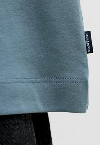 Närbild på fållen av ett ljusblått plagg med dubbelsömnad och en svart tygetikett med texten "JACK & JONES" mot en enfärgad bakgrund.