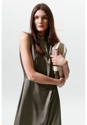 Jeune femme aux longs cheveux bruns portant une robe olive sans manches, tenant un sac à main beige plié sur un fond clair uni.