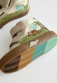 Velcro-toddlerstövlar med en beige canvasöverdel som har dinosaurmotiv; texturerad gummisula i brunt och turkos med zickzackmönster.
