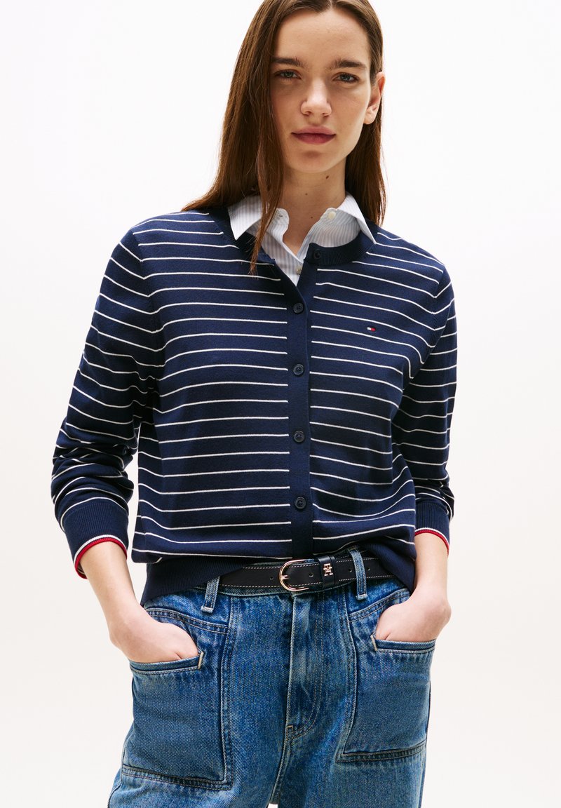 Cardigan a righe navy con chiusura a bottoni, polsini a costine e accento rosso. Indossato sopra una camicia con colletto e abbinato a pantaloni in denim blu.