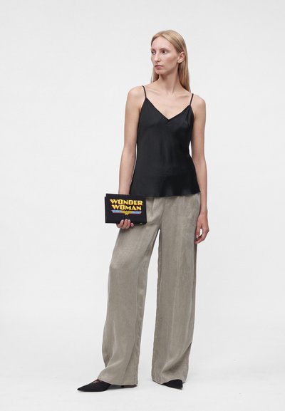 Femme portant un caraco noir et un pantalon beige à jambes larges tient une pochette Wonder Woman, debout devant un fond blanc uni.