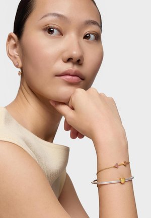 Kobieta w jasnobeżowej bluzce bez rękawów, pozująca z ręką pod brodą, ozdobiona złotymi i srebrnymi bransoletkami w kształcie serca i strzały oraz kolczykami.