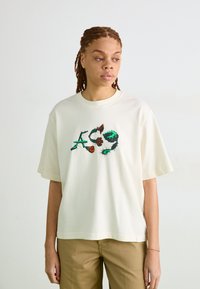 Kremo spalvos per didelis T-shirt, pagamintas iš minkštos medvilnės, su spalvinga raidžių ir formų grafika priekyje, trumpomis rankovėmis, apvaliu kaklu.