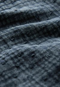 Gros plan sur un tissu foncé texturé avec des bouloches visibles et des bosses douces et en relief, présentant des plis ondulés et une surface légèrement rugueuse.