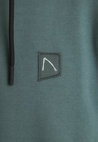 Groene sweatshirt van textured stof. Heeft een zwarte rechthoekige patch met een wit abstract logo en een zwarte trekkoord.