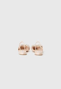 Beige open-back flat sandals with jeweled straps and thin ankle loops, shown from the heel on a plain white background.
