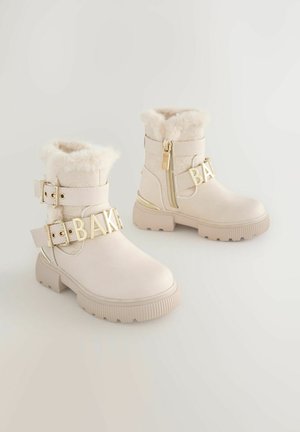 Bottes de cheville couleur crème avec doublure en fausse fourrure, boucles dorées et lettrage décoratif "BAK". Présentées sur un fond clair.