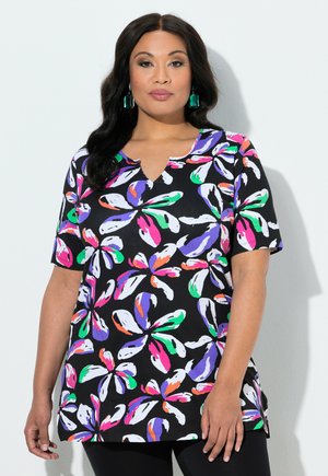 Kvinde med langt mørkt hår iført sort top med korte ærmer og stort farverigt abstrakt blomstermønster samt grønne rektangulære øreringe.