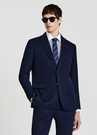 Abito blu navy con tessuto strutturato, due tasche frontali e chiusura con un solo bottone. Abbinato a una cravatta a righe e occhiali da sole.