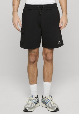 Hombre con pantalones cortos cargo negros con bolsillo lateral, calcetines blancos y zapatillas de correr azul plateado, de pie frente a un fondo gris liso.