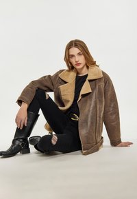 Giacca in shearling marrone foderata, con un ampio colletto, abbinata a jeans neri strappati e stivali neri alti fino al ginocchio. Pelle strutturata e hardware resistente.
