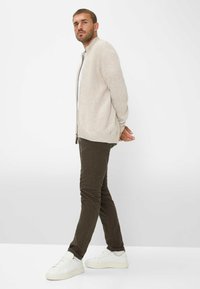 Beige geribbelde cardigan zonder kraag, gecombineerd met donkergroene broek en witte sneakers. Model staat met handen achter de rug, neutrale achtergrond.