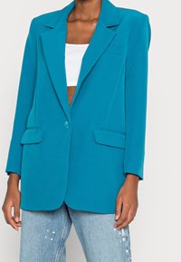 Teal blazer made of lightweight fabric, featuring a notched collar, single button closure, and two front pockets. Worn over a white top.