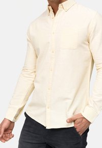Cremefarvet langærmet skjorte med en knapkrave, enkelt brystlomme og tekstureret stof, parret med mørke bukser og en afslappet pasform.