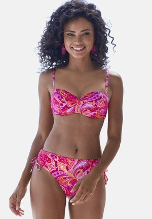 Frau mit lockigem Haar, die einen Bikini mit rosa Paisley-Muster und passende Ohrringe trägt, lächelt und posiert vor einem schlichten Hintergrund.