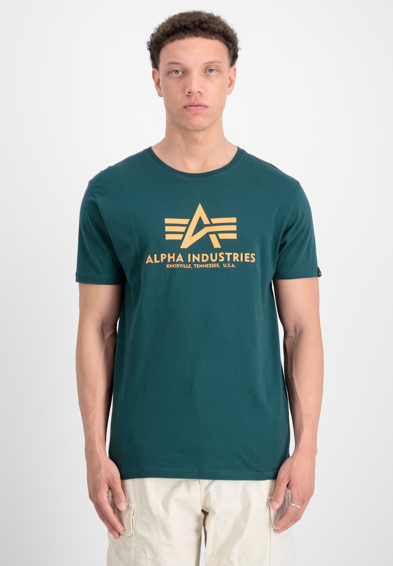 Tyrkis bomulds T-shirt med et markant orange "Alpha Industries" logo med et geometrisk design og korte ærmer, parret med lyse shorts.