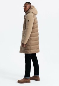 Puffermantel in Hellbraun mit Kapuze, ausgestattet mit gestepptem Futter, Druckknöpfen und einer schlanken Silhouette, kombiniert mit dunklen Jeans und braunen Stiefeln.