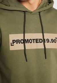 Zelená mikina s kapucí s obdélníkovým béžovým grafickým prvkem s textem "PROMOTED 19,90" v tučném černém písmu. Obsahuje tmavé šňůrky a hladkou texturu.