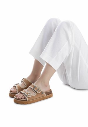 Sandalias planas de ante marrón con tres correas ajustables, costuras decorativas y suela de goma texturizada. Usadas con pantalones blancos cropped.