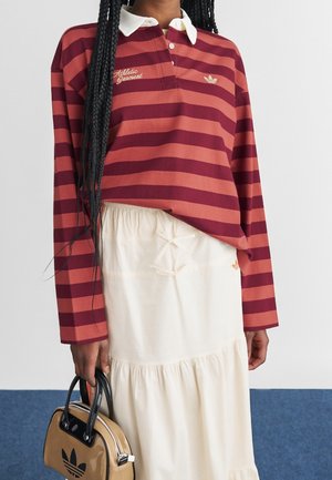 Person trägt ein rot-bordeaux gestreiftes Langarm-Polo und einen cremefarbenen Rock und hält eine beige Adidas-Handtasche mit schwarzen Griffen.