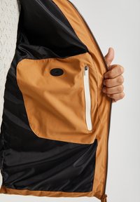 Brązowa podszewka wewnętrzna kurtki z kieszenią na suwak i zaokrąglonym czarnym guzikiem. Materiał jest gładki, z kontrastującą czarną tkaniną.