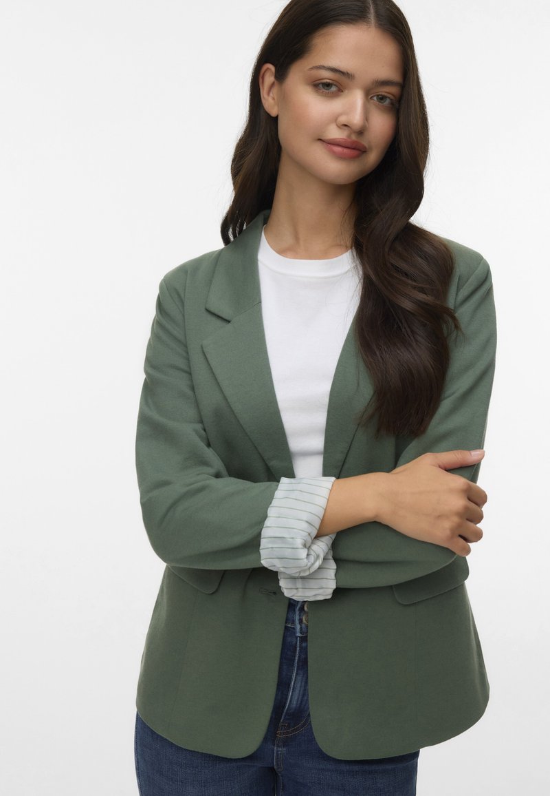 Blazer verde con un design aderente e rever a punta, indossato sopra una camicia bianca con polsini a righe. Abbinato a jeans blu scuro.