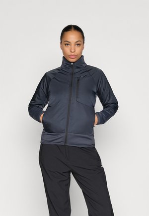 Veste matelassée marine avec un col haut, fermeture éclair à l'avant et une poche poitrine. Présente un design épuré et une texture lisse. Associée avec un pantalon noir.