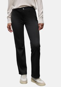 Pantaloni neri realizzati in tessuto liscio, caratterizzati da un design a gamba dritta e una chiusura frontale con bottone. Abbinati a sneaker di colore chiaro.