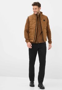 Veste matelassée marron avec col haut, fermeture éclair noire et deux poches avant, portée sur un pull marron texturé et un pantalon noir, assortie à des chaussures noires.