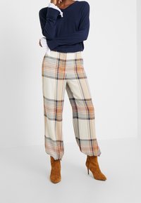 Granatowa bluzka z długim rękawem z falbaniastymi białymi mankietami; kremowe szerokie spodnie w kratę z pomarańczowymi i brązowymi akcentami; brązowe botki na obcasie do kostki.