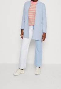 Svetlemodrý kabát s moderním siluetou, spárovaný s vícebarevným pruhovaným svetrem, bílými džíny a béžovými teniskami.