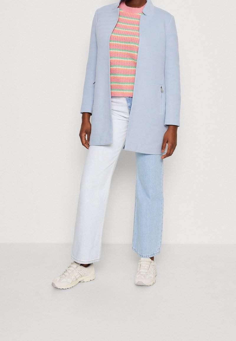 Svetlemodrý kabát s moderním siluetou, spárovaný s vícebarevným pruhovaným svetrem, bílými džíny a béžovými teniskami.