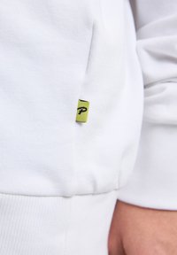 Felpa bianca con una texture morbida, dotata di polsini a coste e un piccolo etichetta verde con logo nero cucita sulla cucitura.