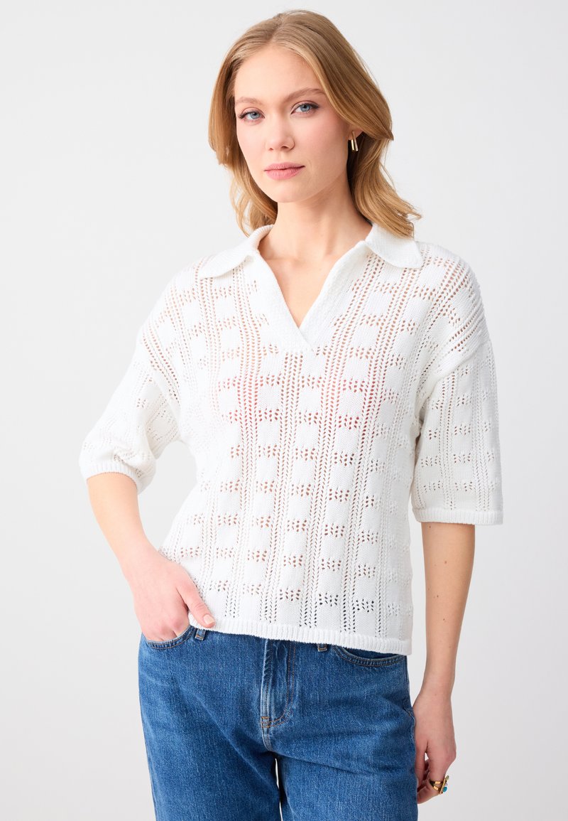 Femme blonde portant un pull blanc à col en V en maille ajourée avec col et un jean bleu, posant avec une main dans la poche sur un fond uni.