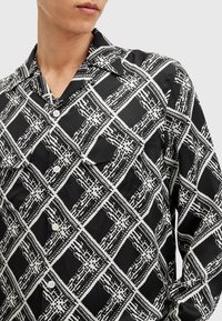 Camicia a righe bianco e nero con un design geometrico e trame di linee diverse, maniche lunghe e colletto classico. Due bottoni visibili.