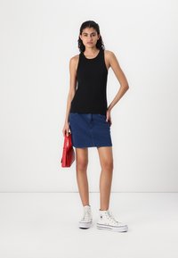 Zwarte mouwloze tanktop, denim rok, rode handtas en witte hoge sneakers op een model tegen een effen achtergrond.