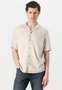 Camisa beige de manga corta con un sutil patrón de remolino blanco, cuello abotonado y botones marrones, combinada con unos vaqueros azul oscuro.